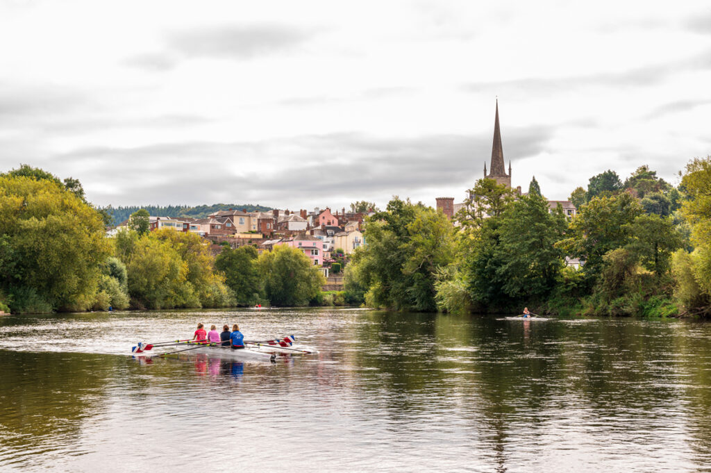 A view of Ross-On-Wye town from the river. A group of people rowing on the river and buildings and a church spire is on the horizon.